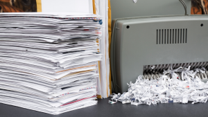 A stack of papers sits beside a gray paper shredder on a table. Shredded paper strips are visible, indicating document destruction. The scene conveys efficiency and organization.
