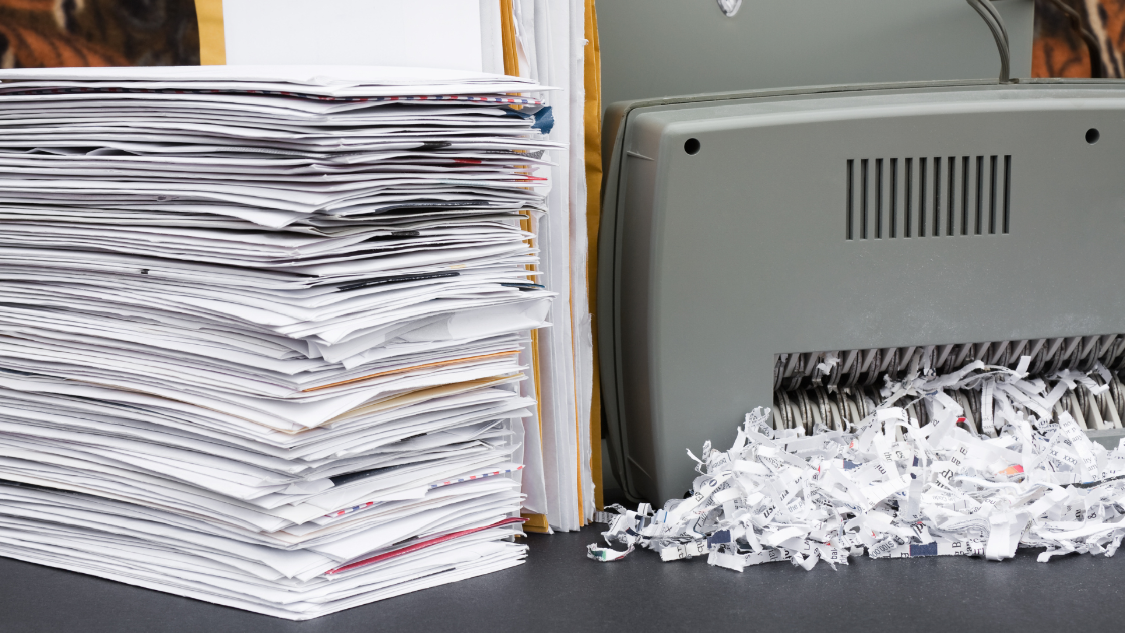 A stack of papers sits beside a gray paper shredder on a table. Shredded paper strips are visible, indicating document destruction. The scene conveys efficiency and organization.